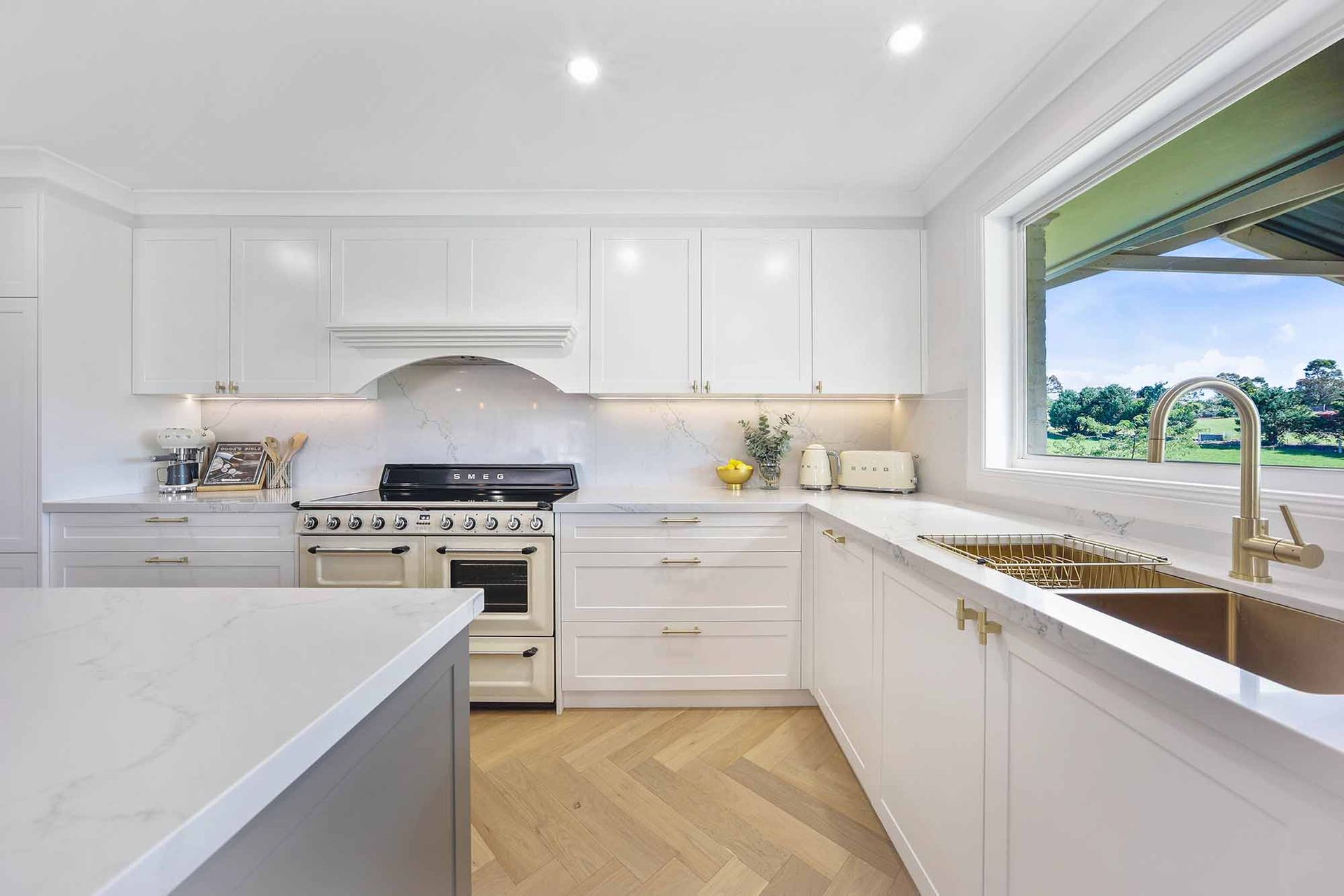 White kitchen with stainless steel appliances, gold fixtures, and a large window overlooking a green landscape — Above & Beyond Interiors Custom Joinery in Bellambi, NSW