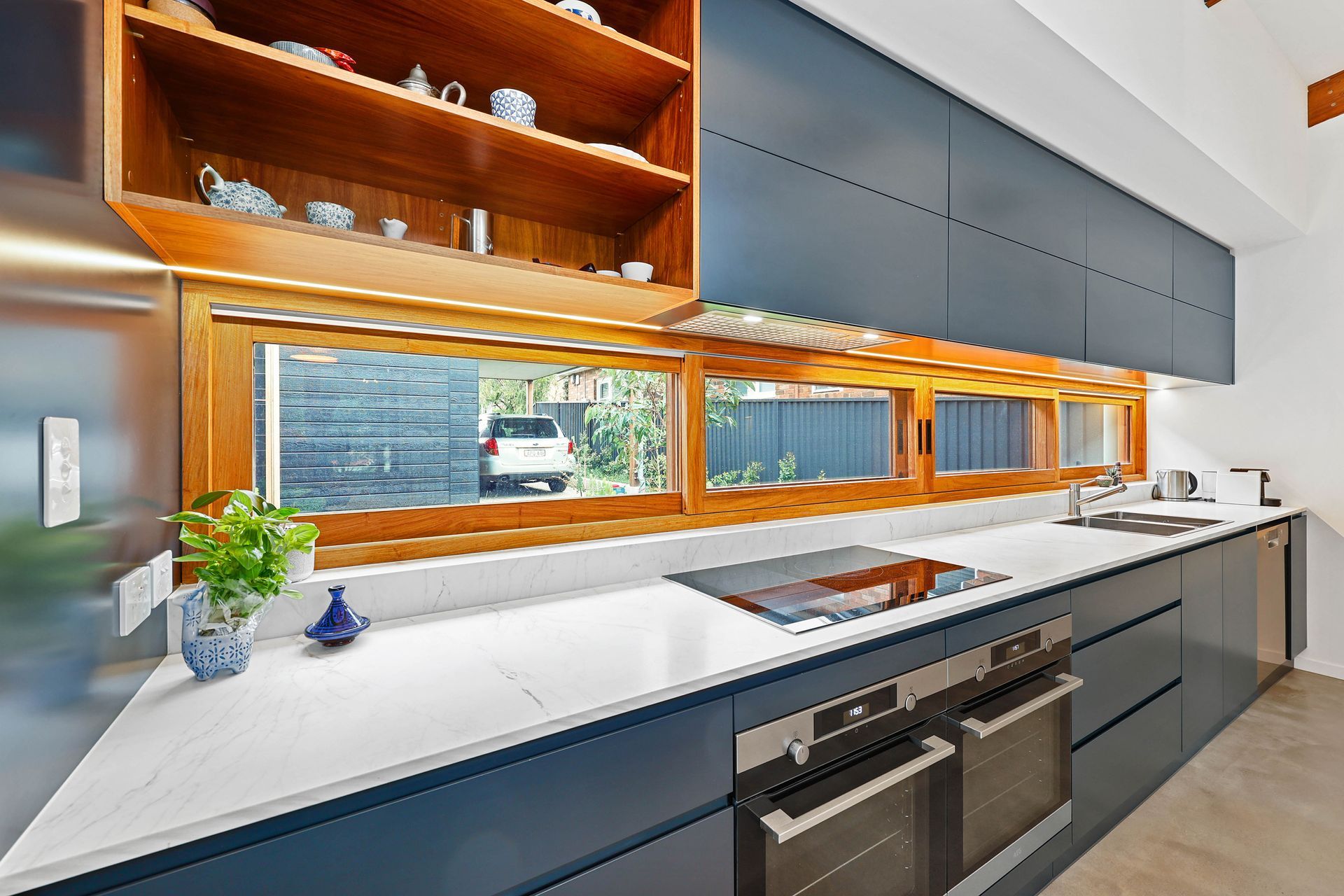 Modern kitchen with blue cabinets, wooden shelves, white countertops, and a window overlooking a car — Above & Beyond Interiors Custom Joinery in Bellambi, NSW