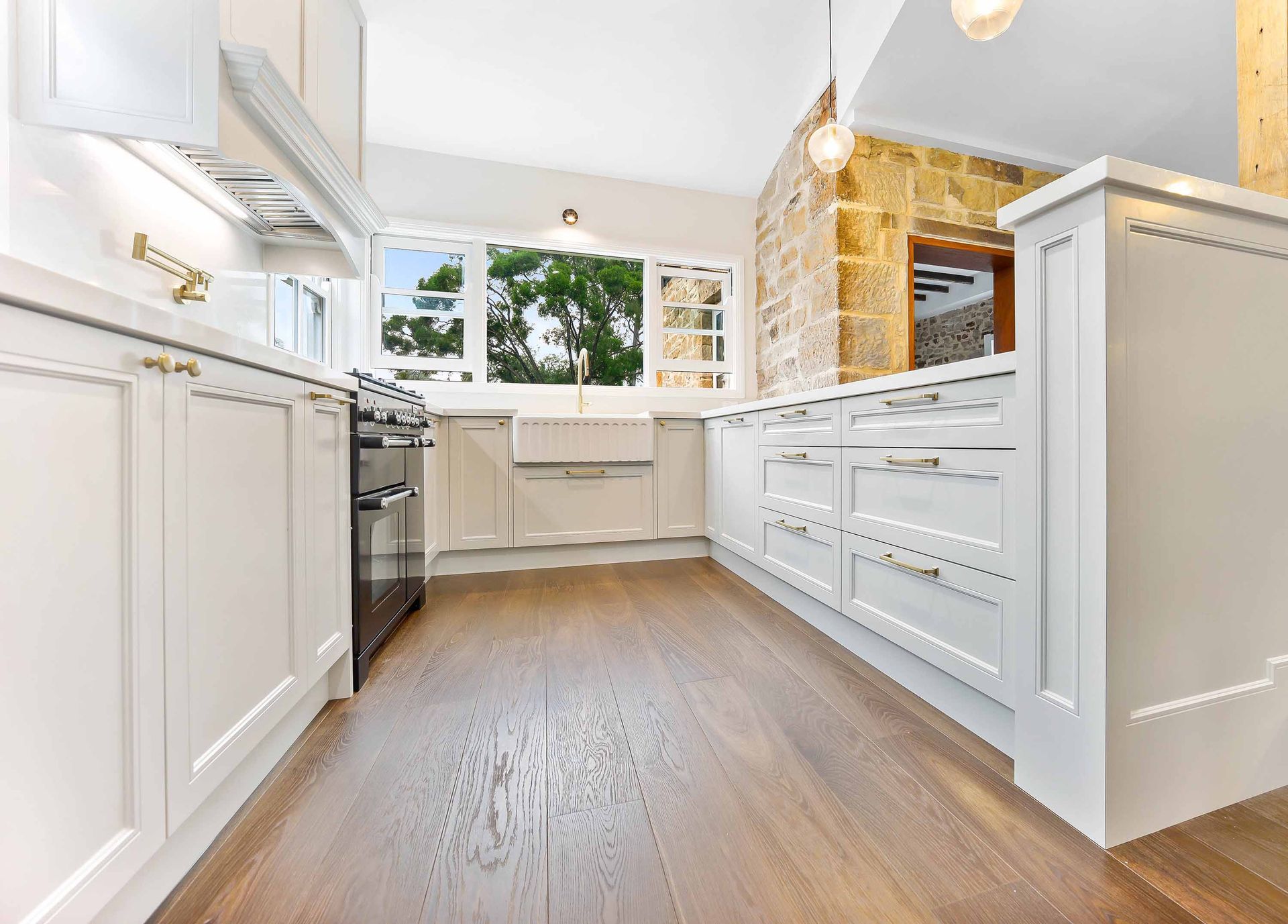 White kitchen with wooden floors, cabinetry, and stone wall. Window views greenery — Above & Beyond Interiors Custom Joinery in Bradbury, NSW
