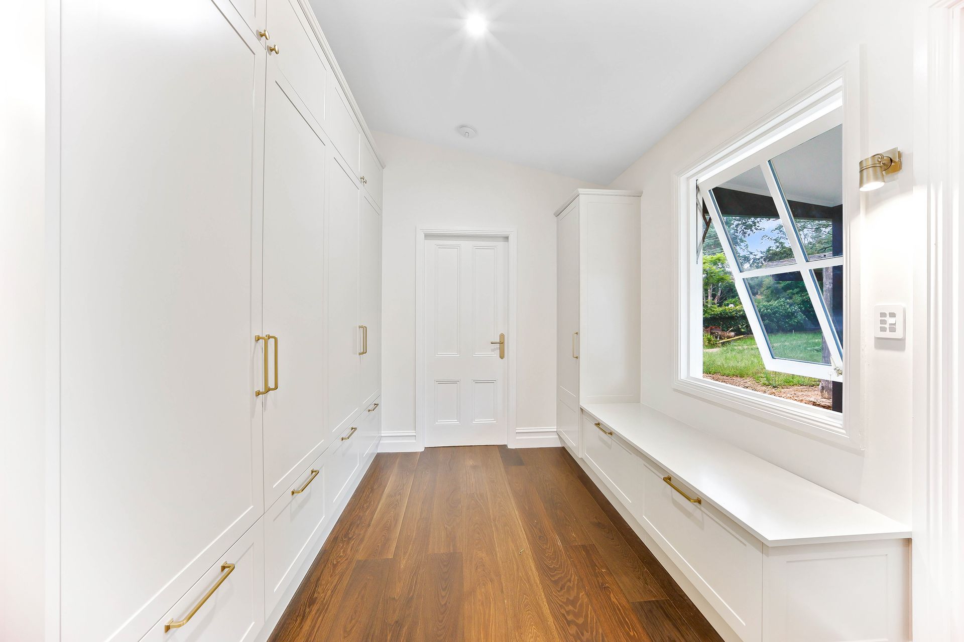 Bright white hallway with built-in cabinets, wooden floor, and window with bench seat — Above & Beyond Interiors Custom Joinery in Bradbury, NSW