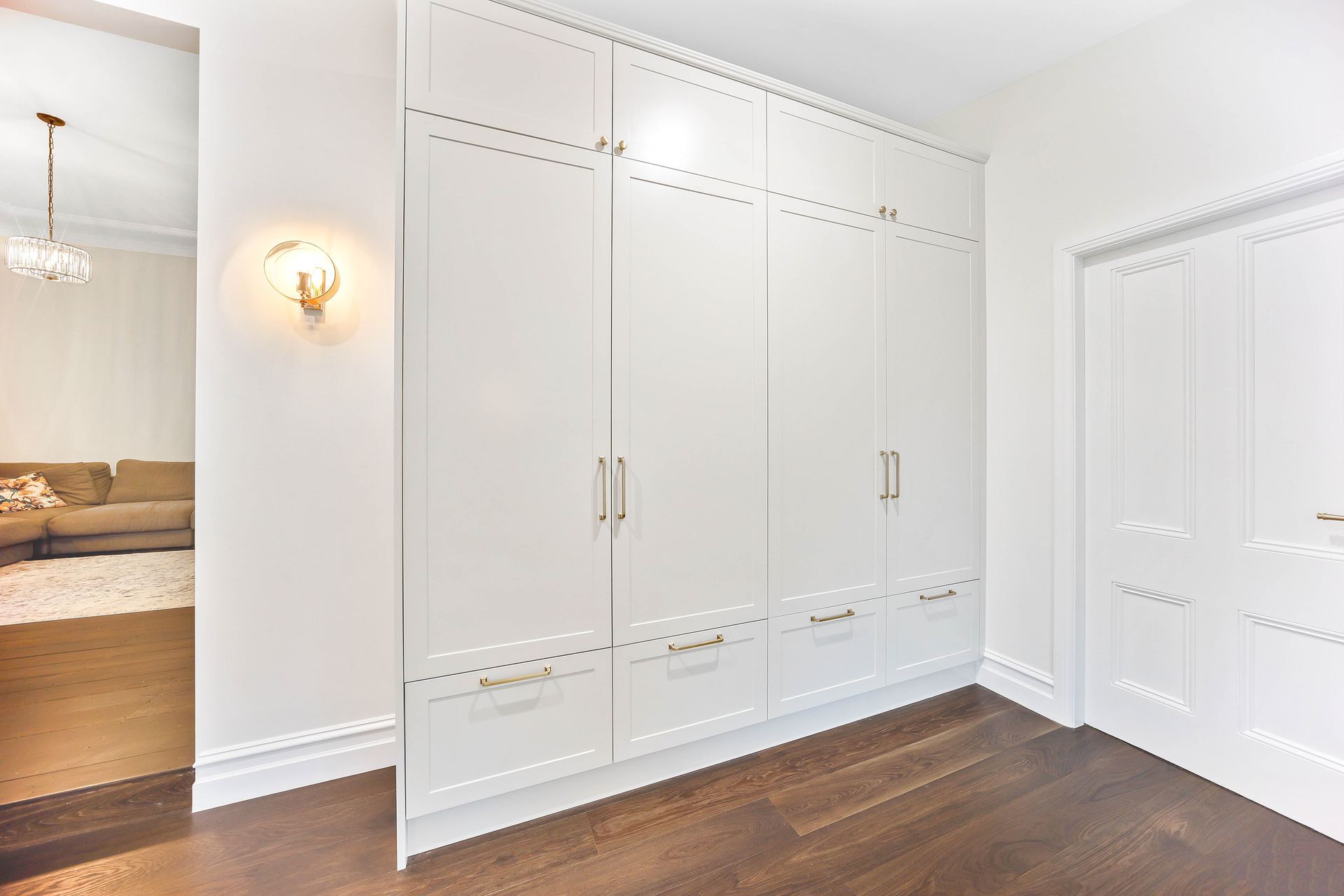White built-in closet with gold hardware and a white door in a hallway with dark wood floors — Above & Beyond Interiors Custom Joinery in Bradbury, NSW