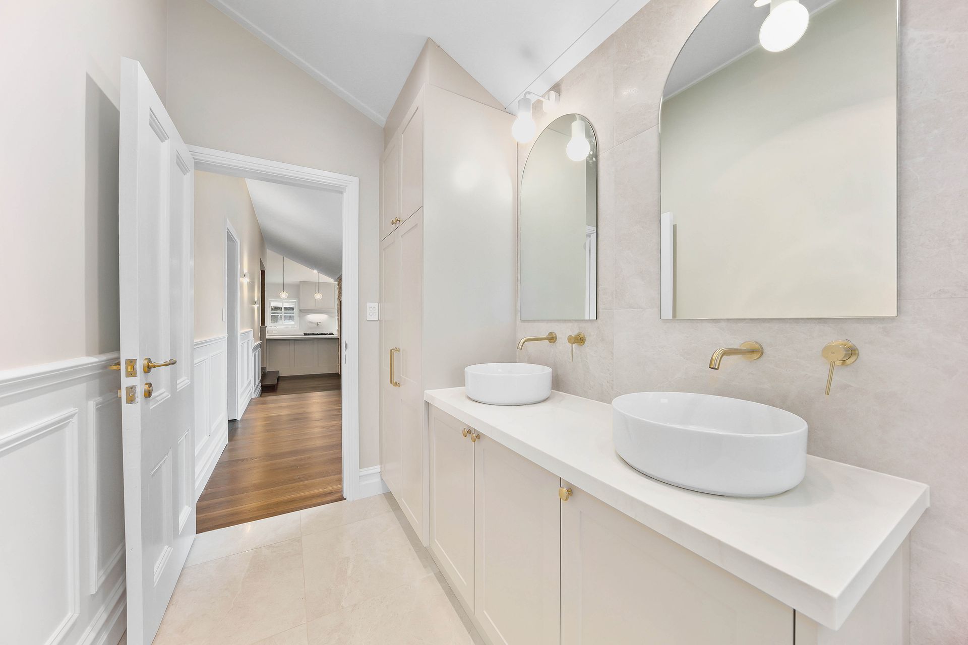 A modern bathroom with white cabinets, sinks, and gold fixtures. Doorway leads to a hallway — Above & Beyond Interiors Custom Joinery in Bradbury, NSW