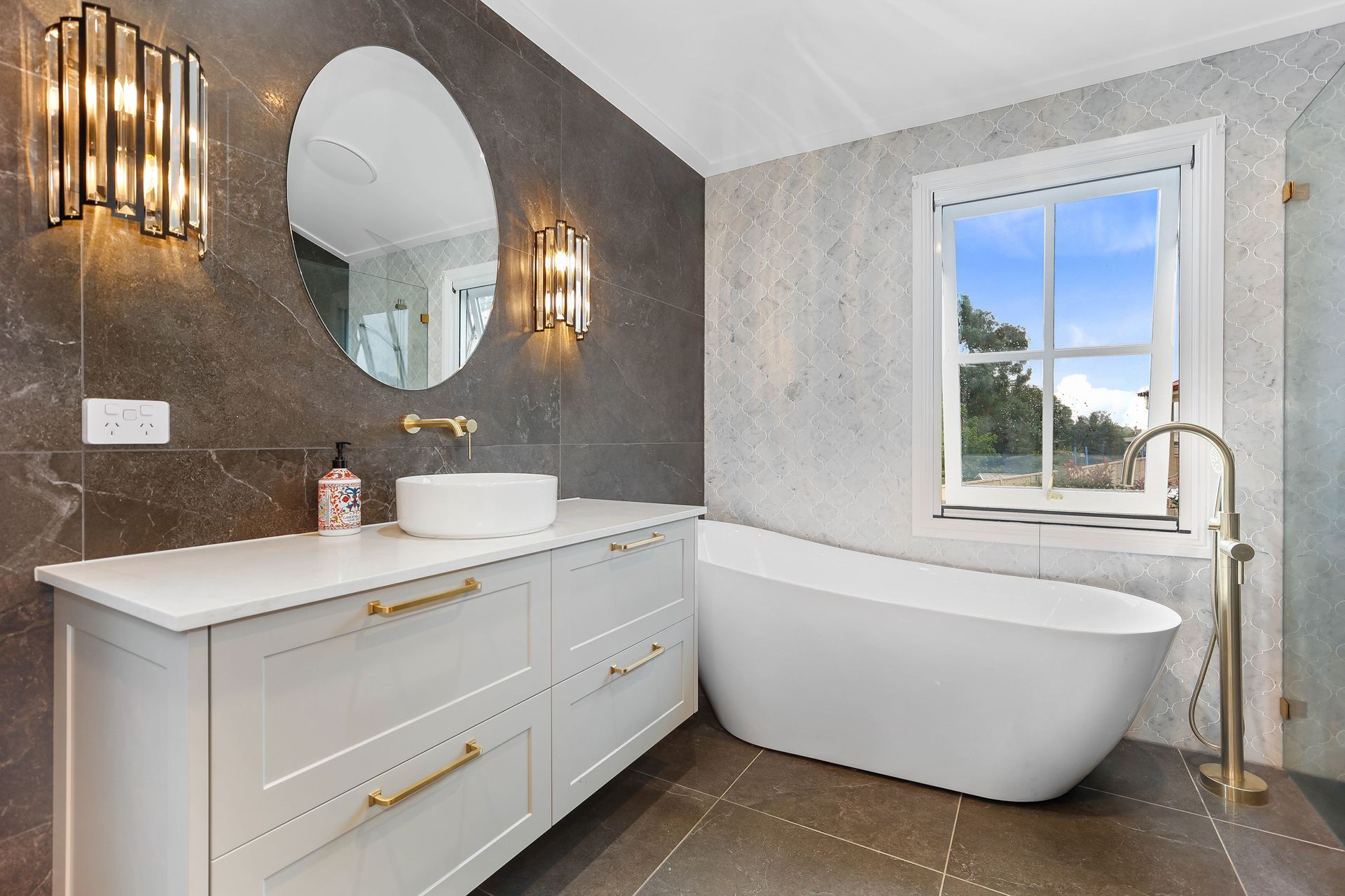 Modern bathroom with white vanity, gold fixtures, grey tiled walls, and a freestanding bathtub — Above & Beyond Interiors Custom Joinery in Bradbury, NSW