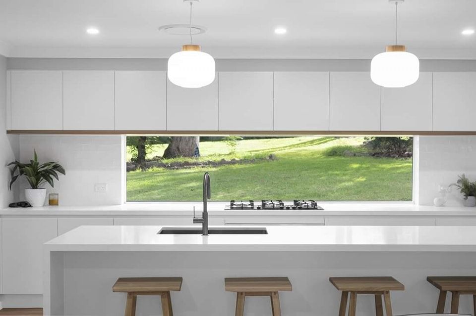 Modern white kitchen with island, wooden stools, and a long window overlooking a green lawn. Two pendant lights hang above — Above & Beyond Interiors Kitchen Renovation in Smeaton Grange, NSW