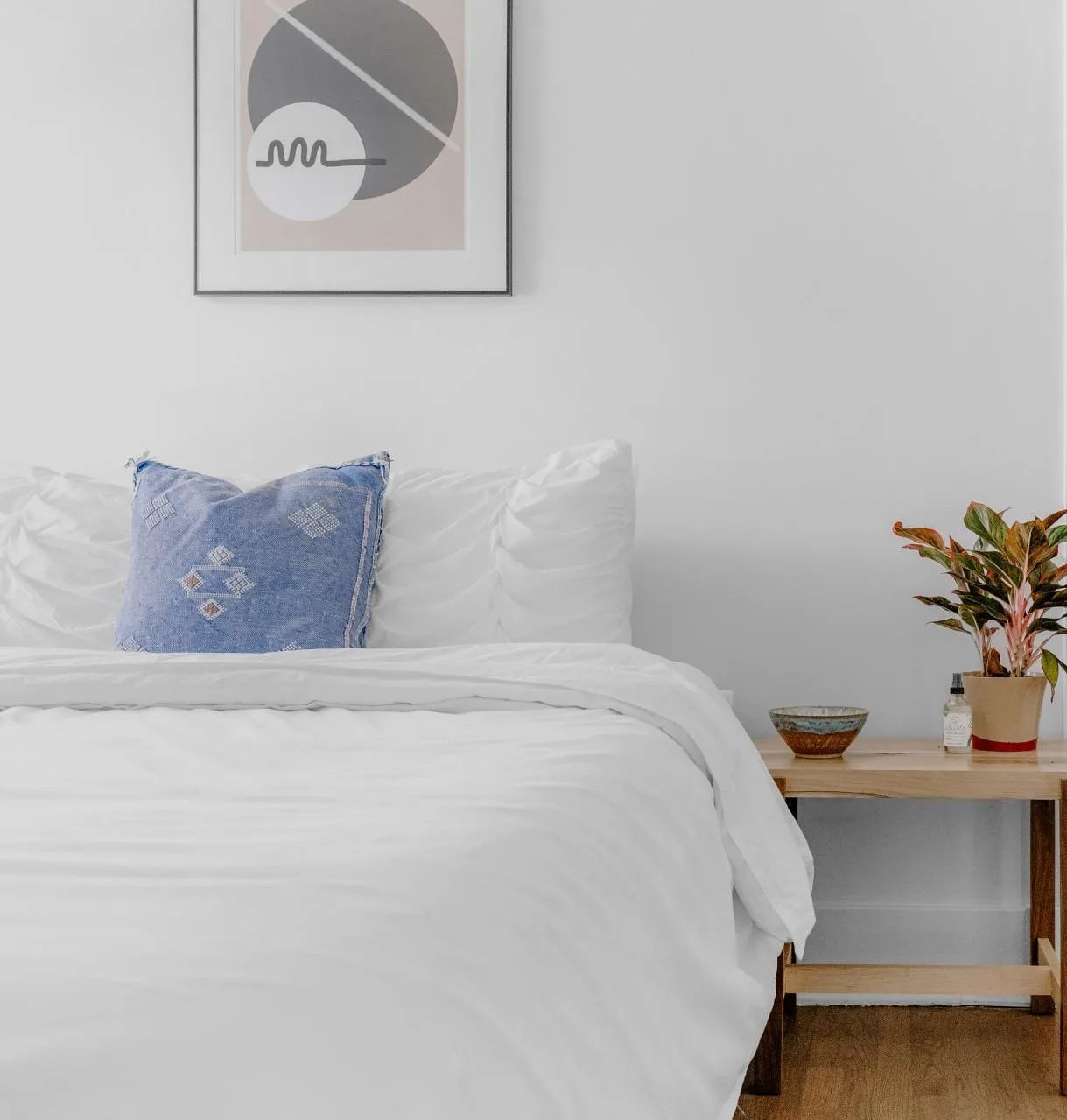 Bedroom with white bedding, blue pillow, wooden side table, and art.