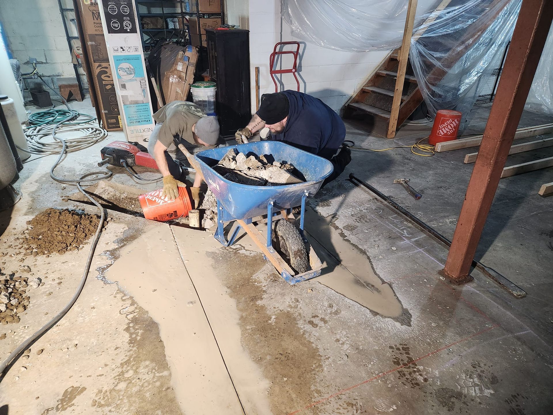 Two men are working in a basement with a wheelbarrow full of dirt.