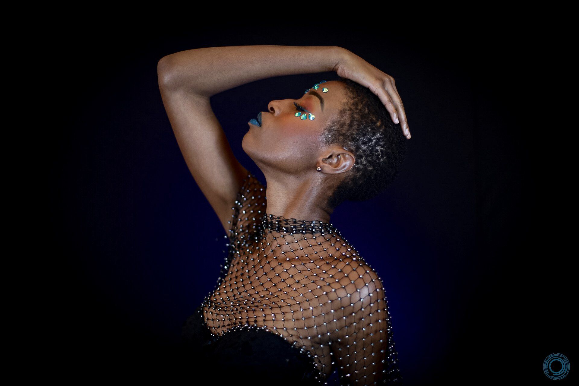 A woman in a black dress with rhinestones on her face is standing with her hand on her head.