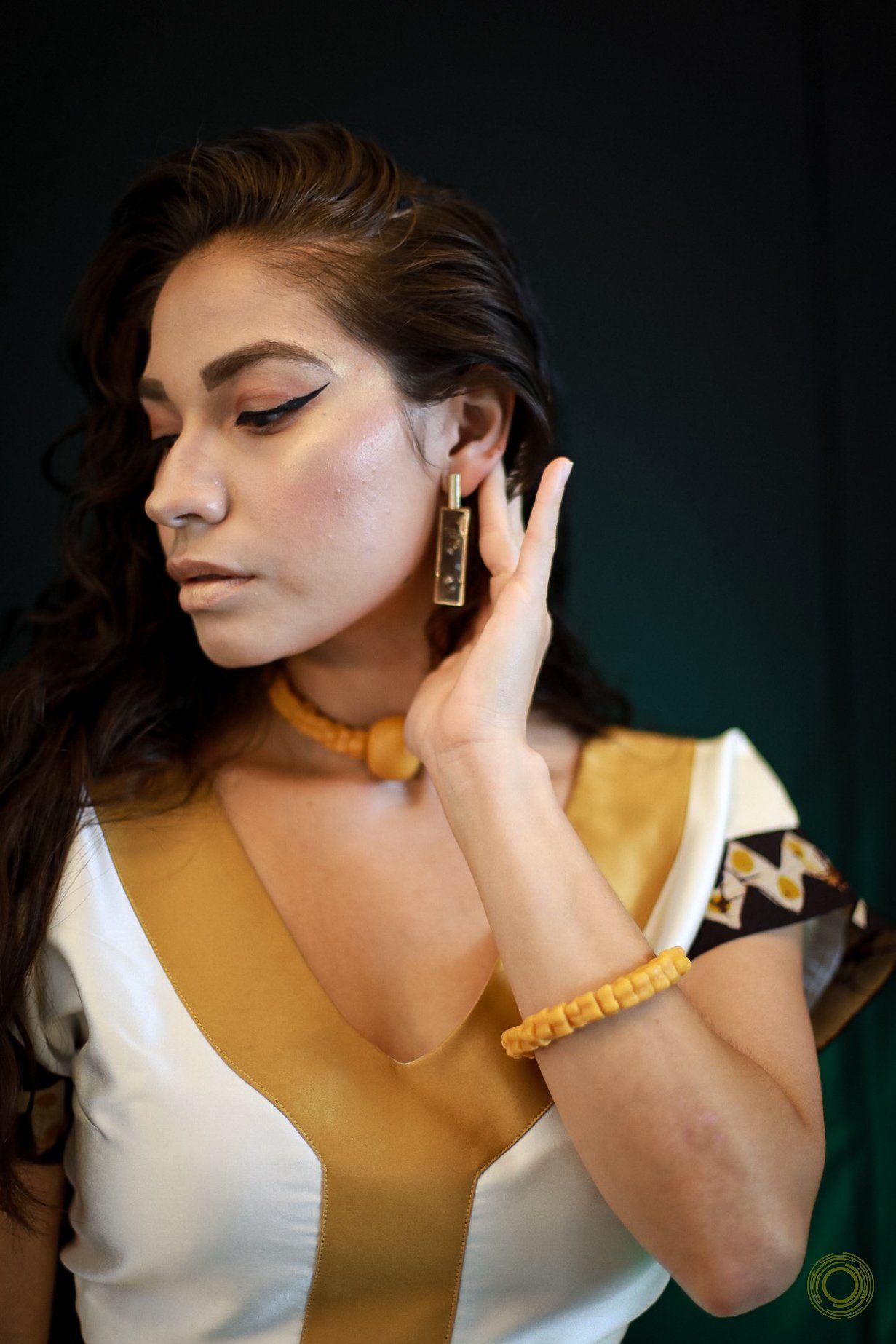A woman wearing a white and gold dress and earrings is touching her ear.