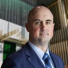 Man in blue suit, smiling slightly, in a modern building with glass and wood.