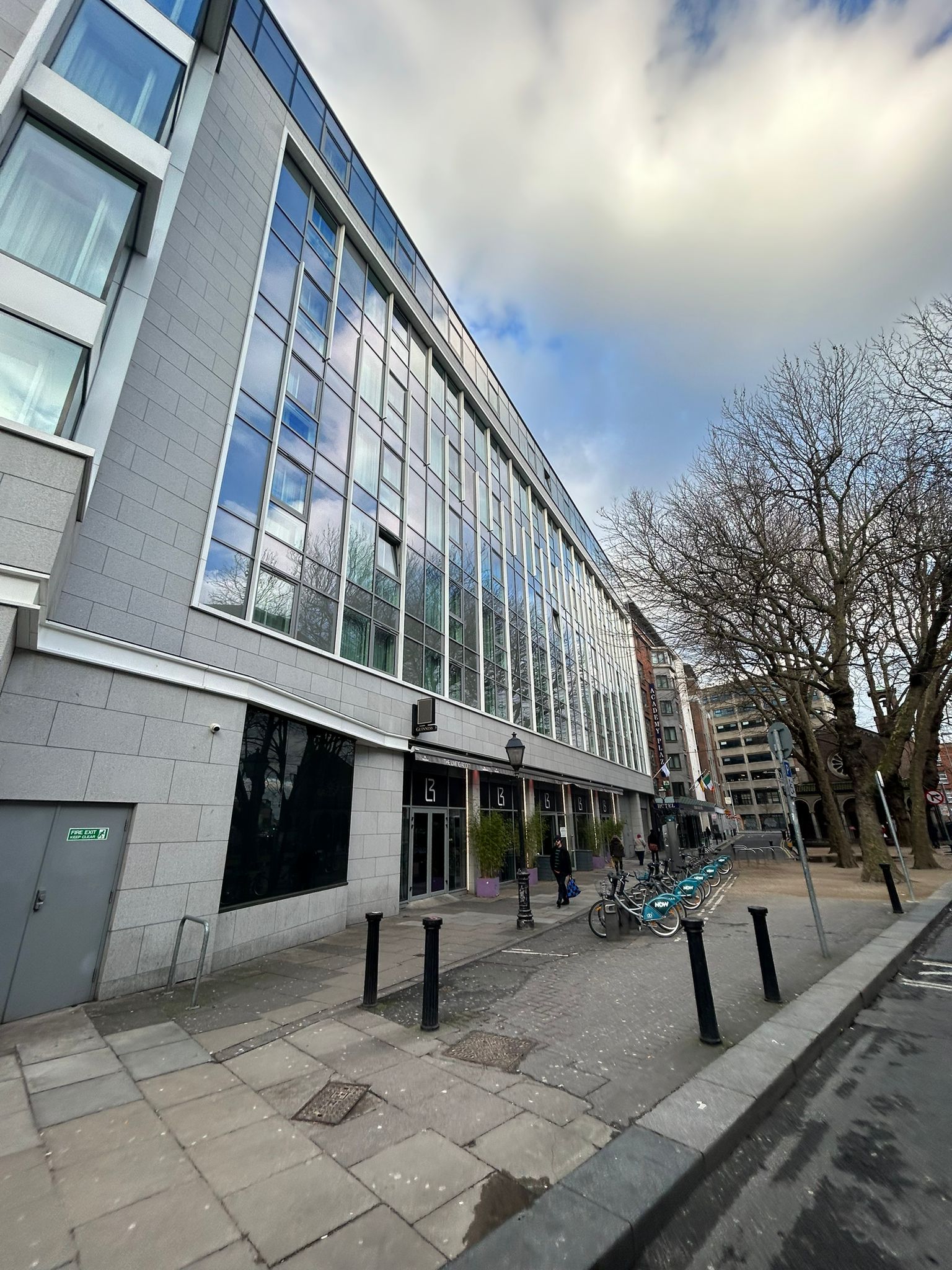 A large building with a lot of windows is surrounded by trees and a sidewalk.