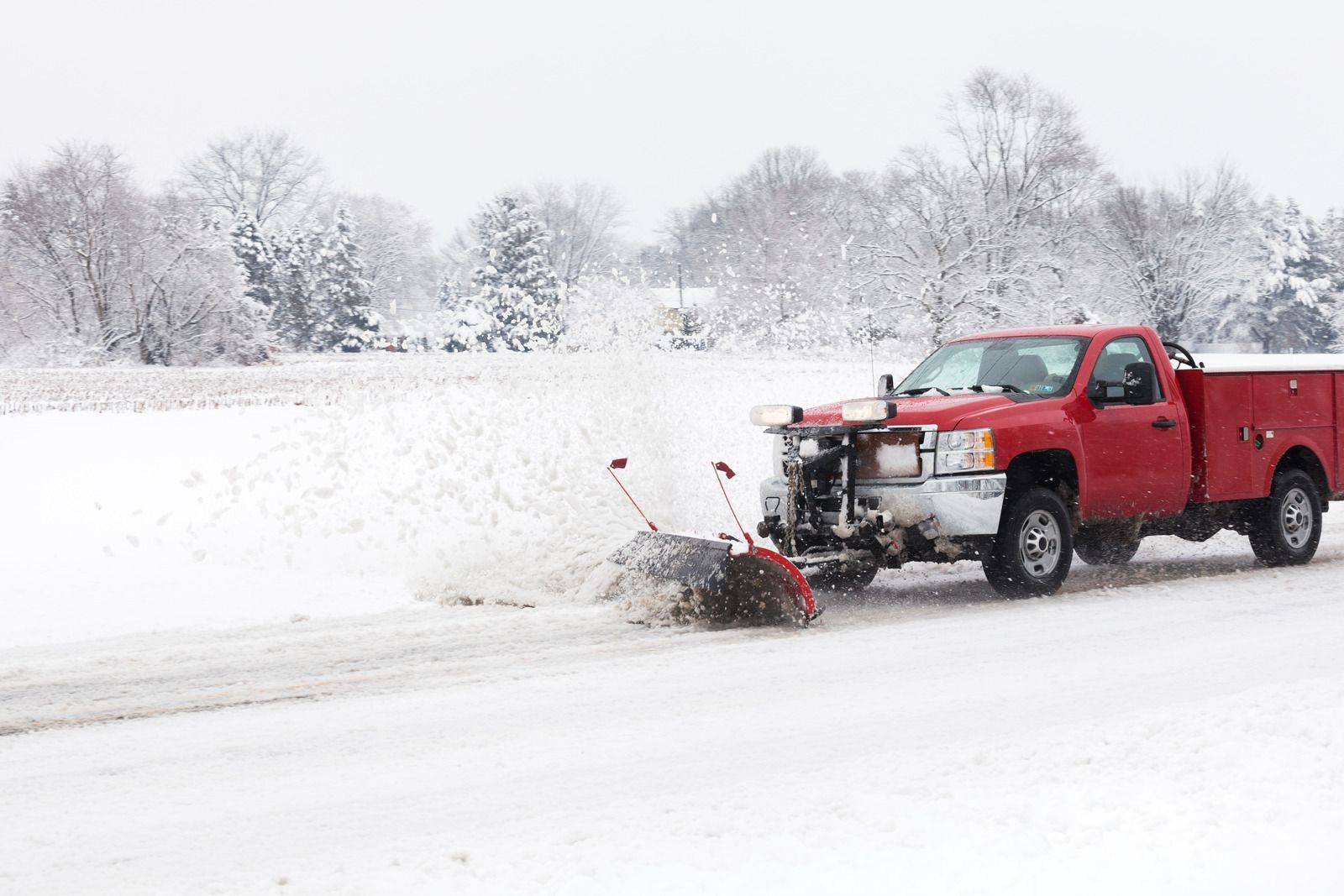 A red pickup truck with a snowplow blade attached moves through a snowy field, spraying snow as it clears the ground.