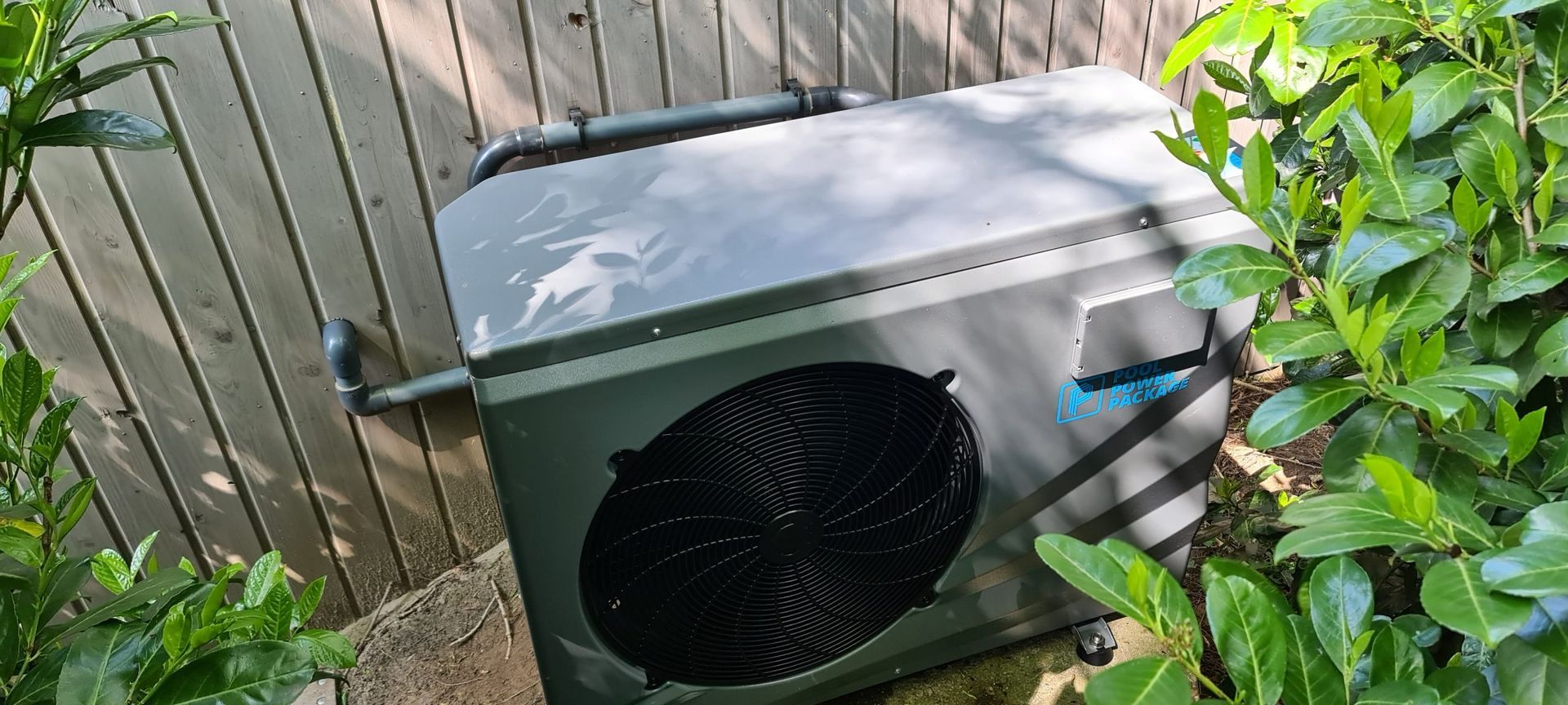 Een zwarte airconditioner staat op de grond naast een houten hek.