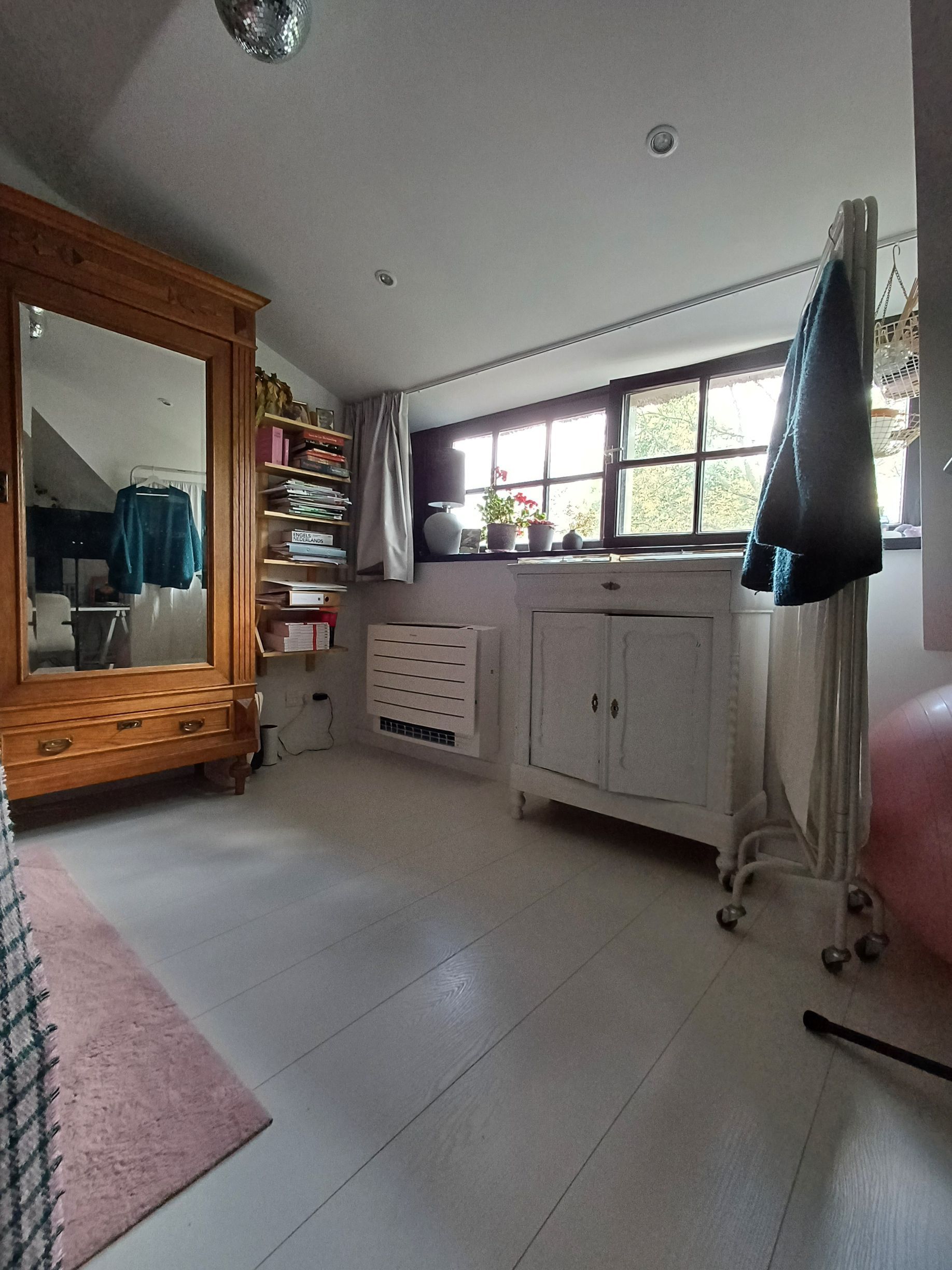 Sfeervolle zolderkamer met wit houten vloer, houten kast, boekenplanken en een Daikin Perfera vloermodel airco onder het raam voor efficiënte klimaatregeling.