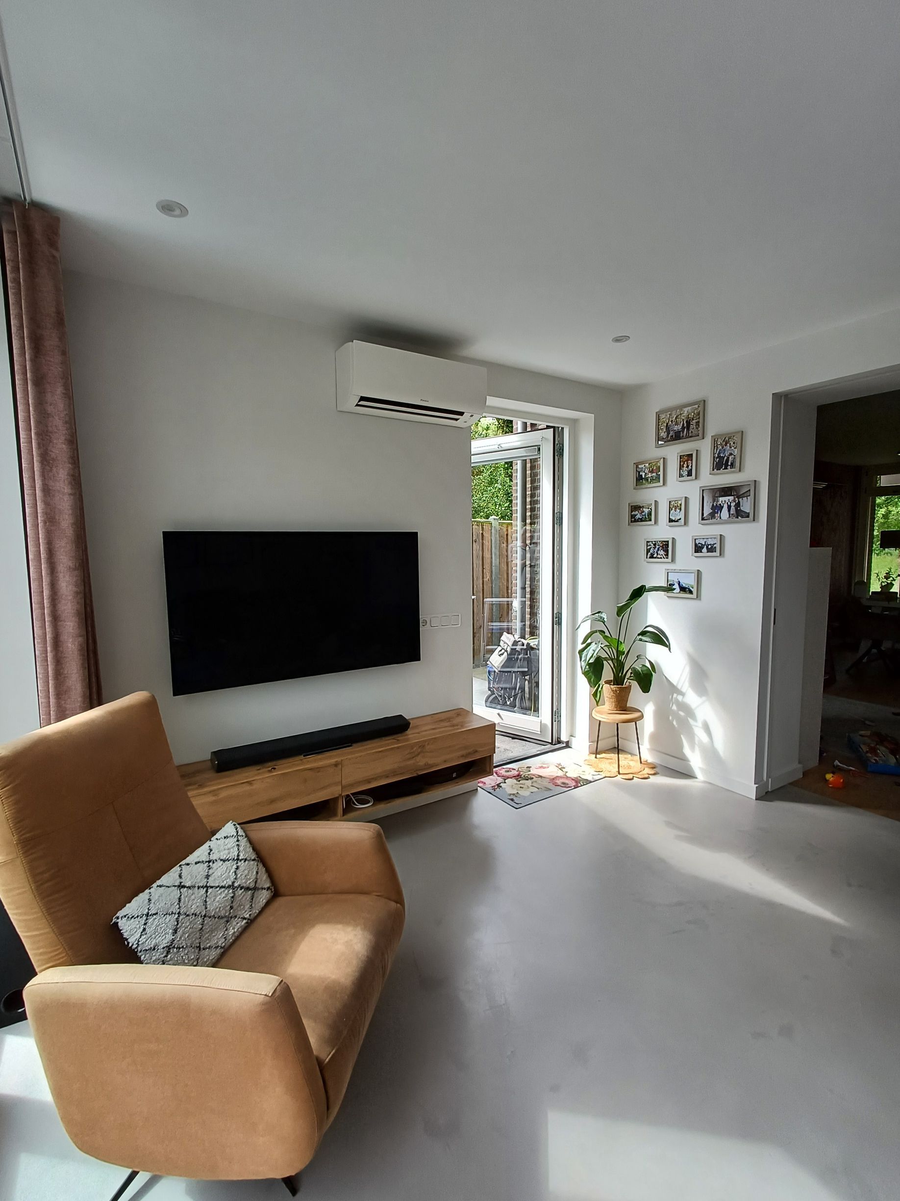 Daikin Comfora airco strak gemonteerd boven de televisie in een lichte, moderne woonkamer met houten tv-meubel, fauteuil en grote raampartij voor natuurlijk licht.