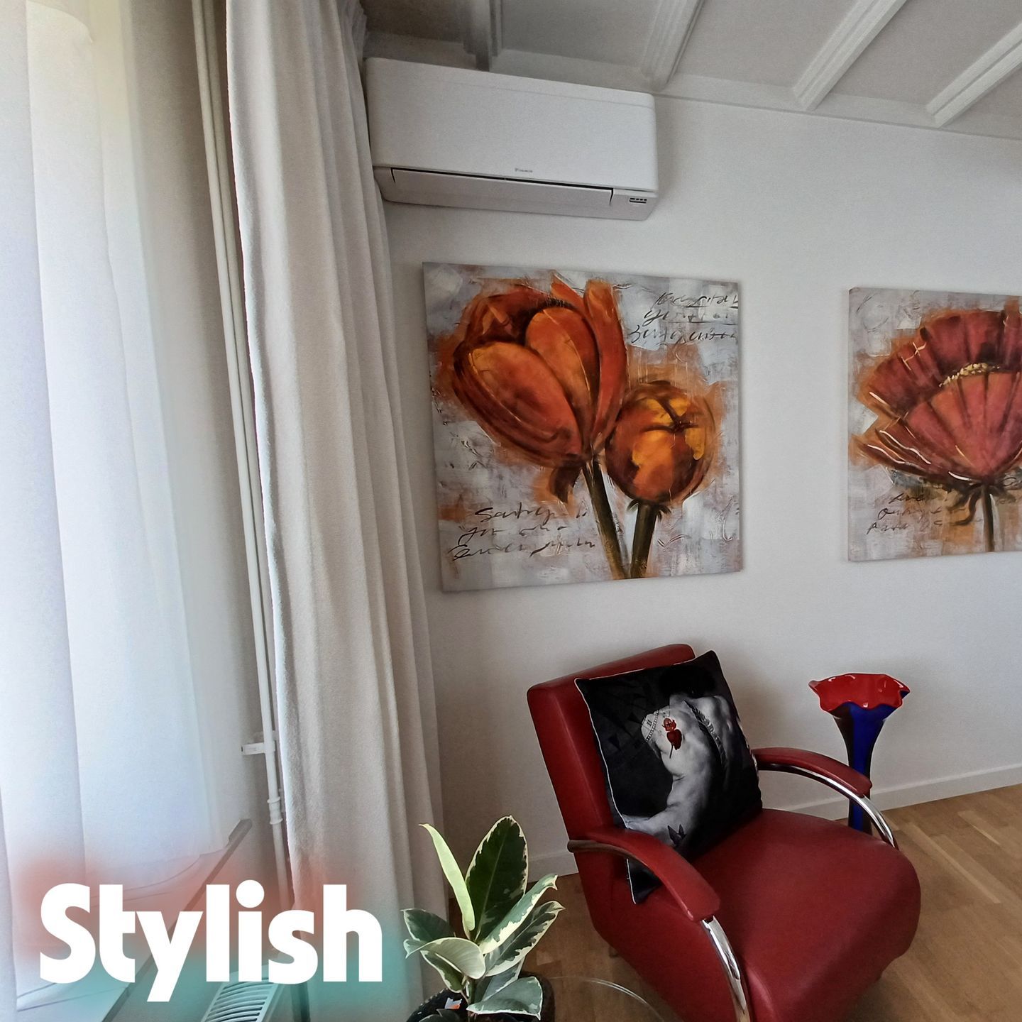 Woonkamerhoek met een witte Daikin Stylish airco aan de muur boven twee kleurrijke bloemenschilderijen, een rode designstoel met een kussen en een plant op de voorgrond, met houten vloer en modern interieur.