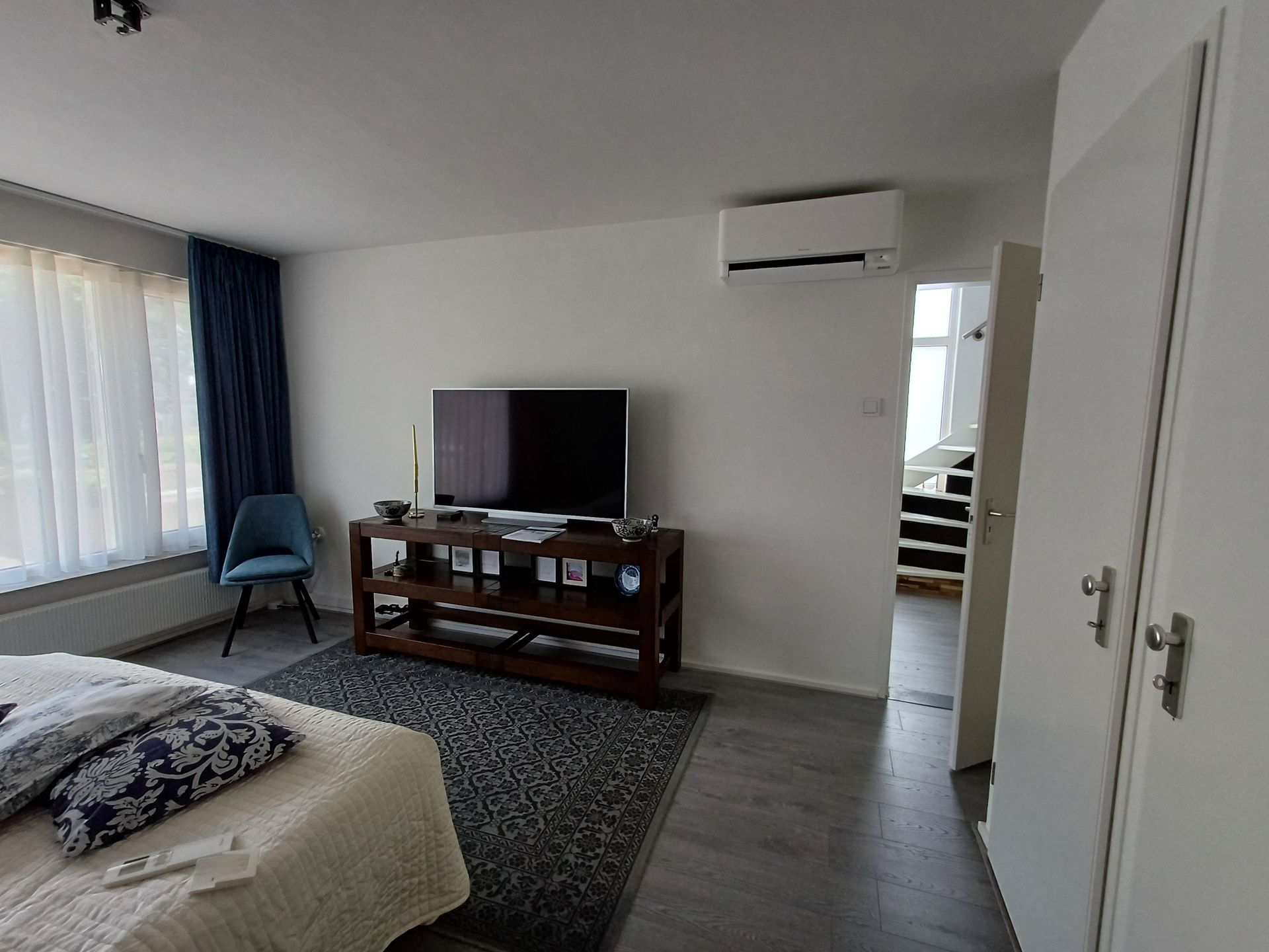 Slaapkamer met een witte Daikin Stylish airco aan de muur, een televisie op een donkerhouten meubel, een bed met sierkussens, een blauw gordijn en een deur die uitzicht geeft op een trap naar boven.