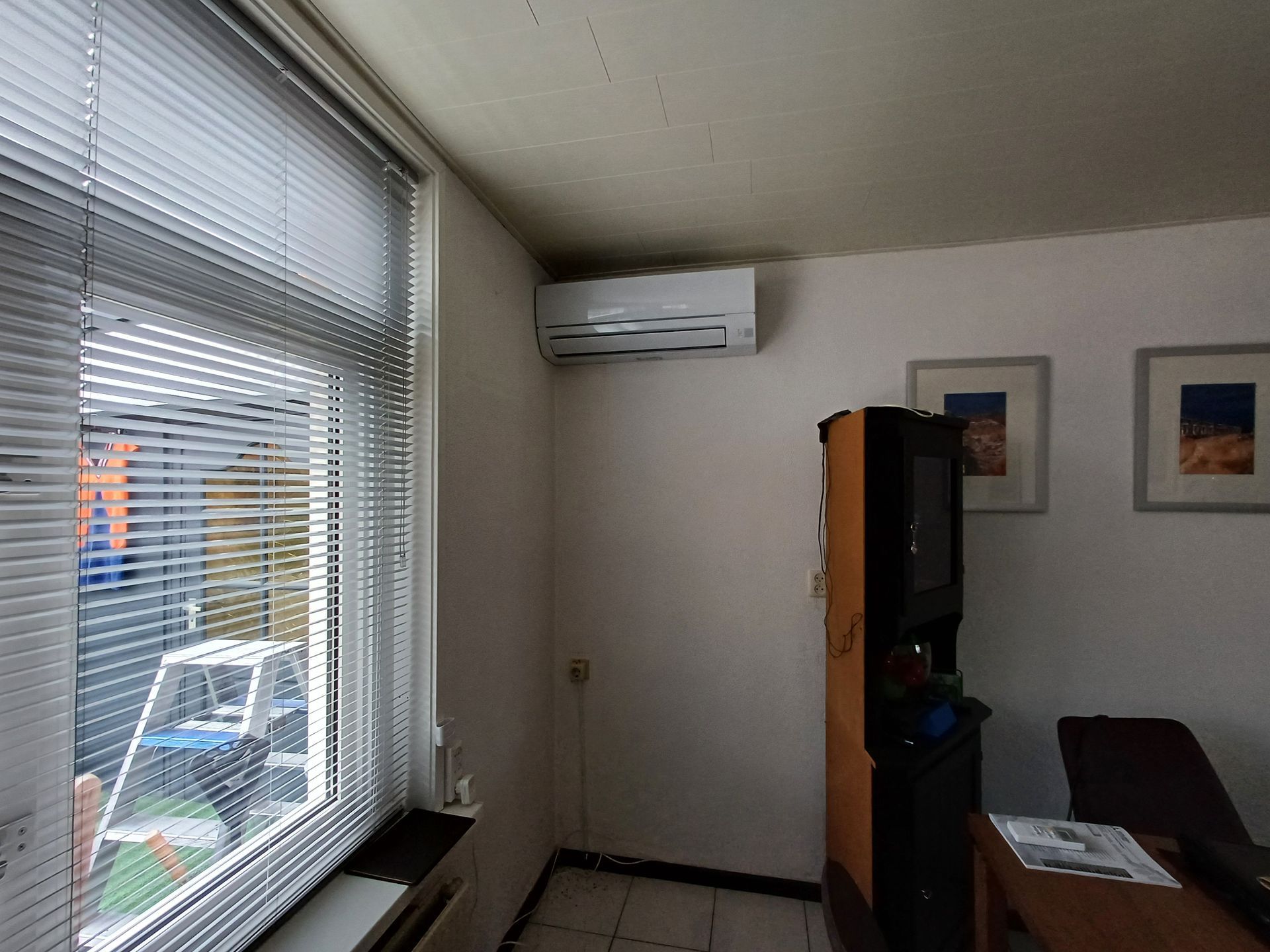 Kleine kamer met een witte Mitsubishi Electric Basis airco aan de muur, houten kast eronder, jaloezieën voor het raam en een tafel met papieren op de voorgrond.