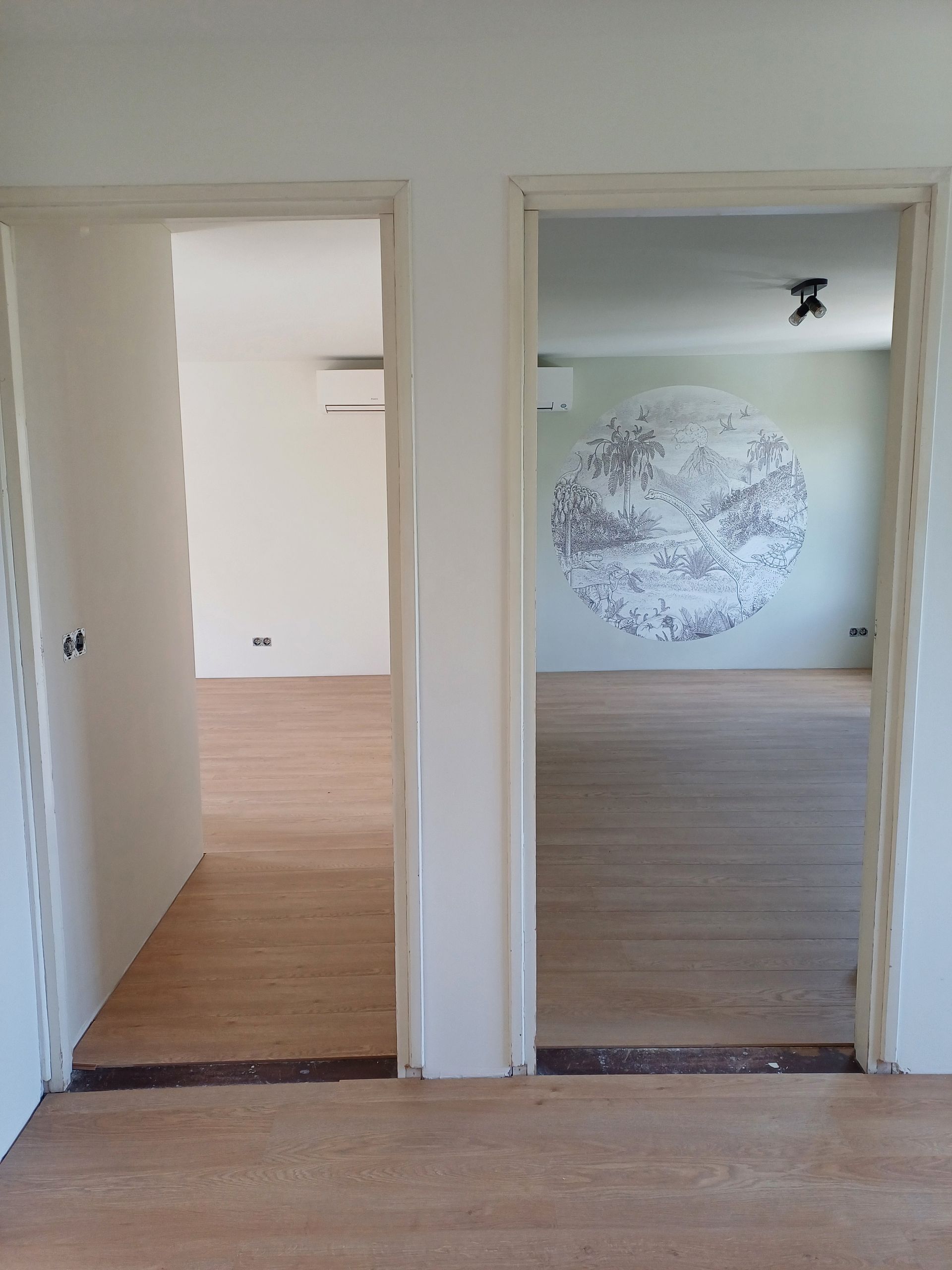 Twee naast elkaar gelegen kamers met houten vloer, elk voorzien van een witte Daikin Perfera airco aan de muur; in de rechterkamer is een groot rond wandkunstwerk met een tropisch landschap zichtbaar.