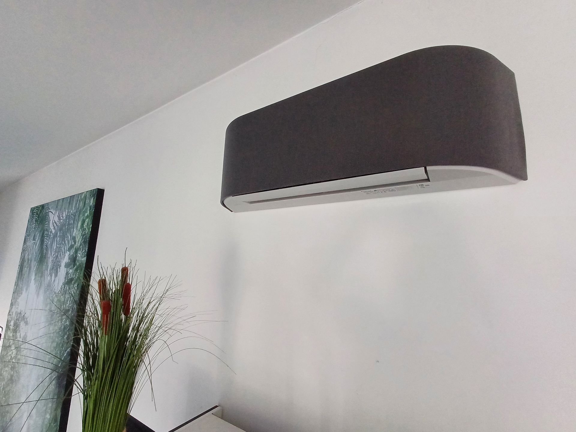 Toshiba Haori airco in donkergrijze stofafwerking gemonteerd aan een witte muur, met decoratieve planten en een schilderij op de achtergrond.