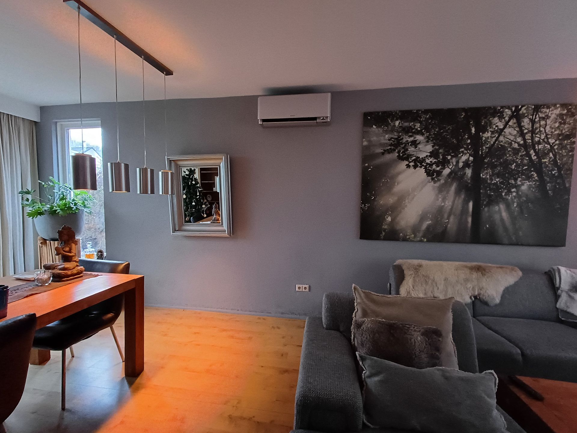 Stijlvolle Daikin Stylish airco voor comfortabele klimaatbeheersing in de woonkamer. Moderne woonkamer met grijze muren, houten vloer, een Daikin Stylish airco aan de muur, een grote zwart-wit wandfoto van een bos, een grijze hoekbank en een houten eettafel met hanglampen en decoraties.