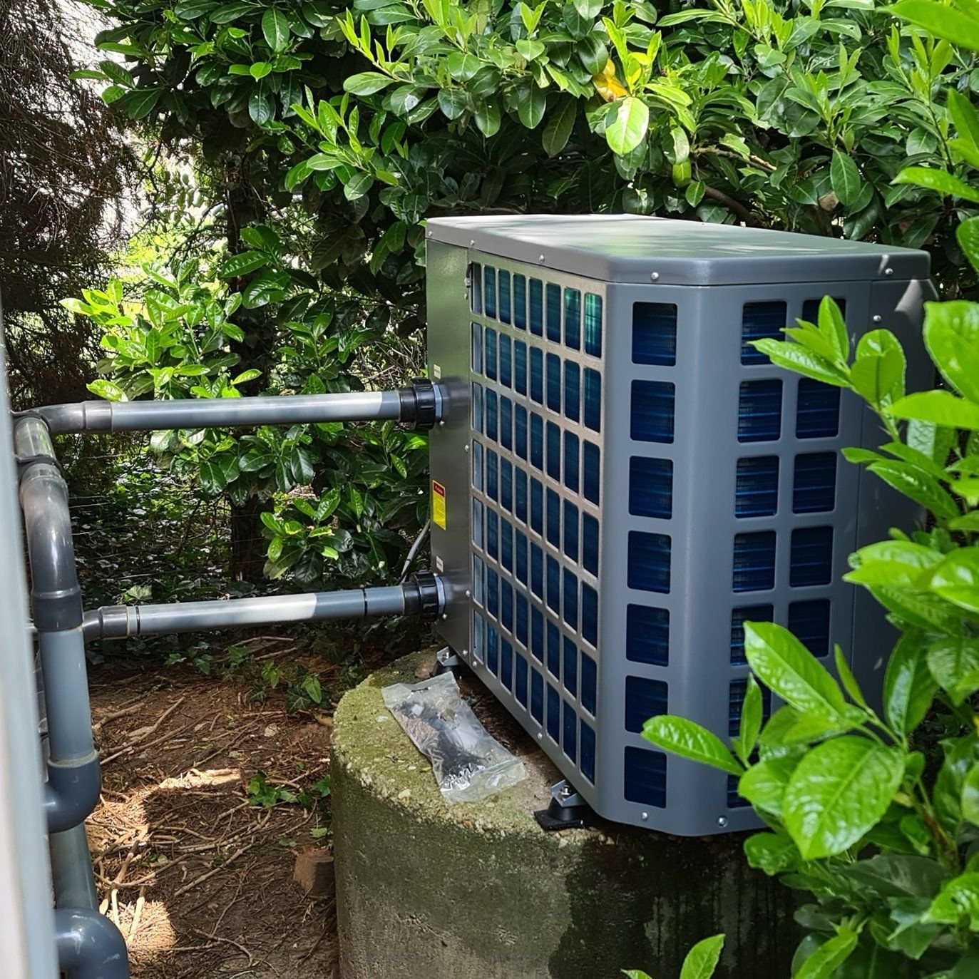 Een grijze airconditioner staat op een betonnen blok, omringd door bomen.