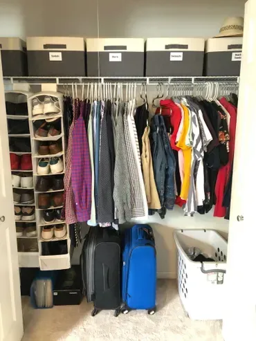 Closet with clothes on hangers, shoe organizer, boxes on top shelf, suitcases, and laundry basket.