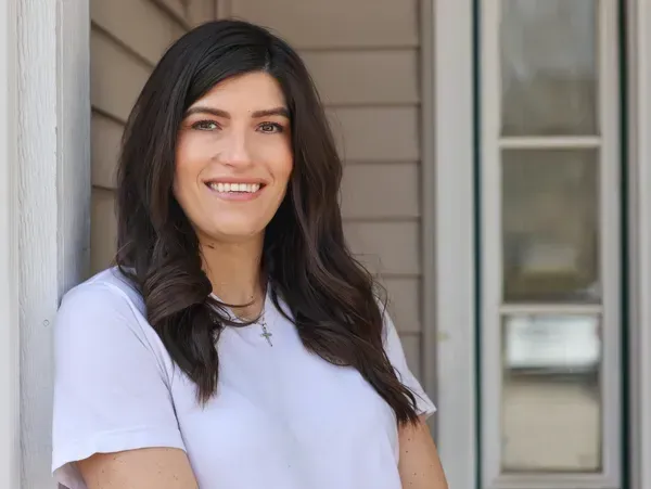 Woman with long dark hair smiles, leaning against a white doorframe. She wears a white shirt.