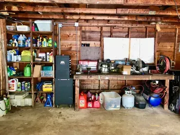 Cluttered garage interior with wooden shelves, workbench, and various tools, equipment, and supplies.
