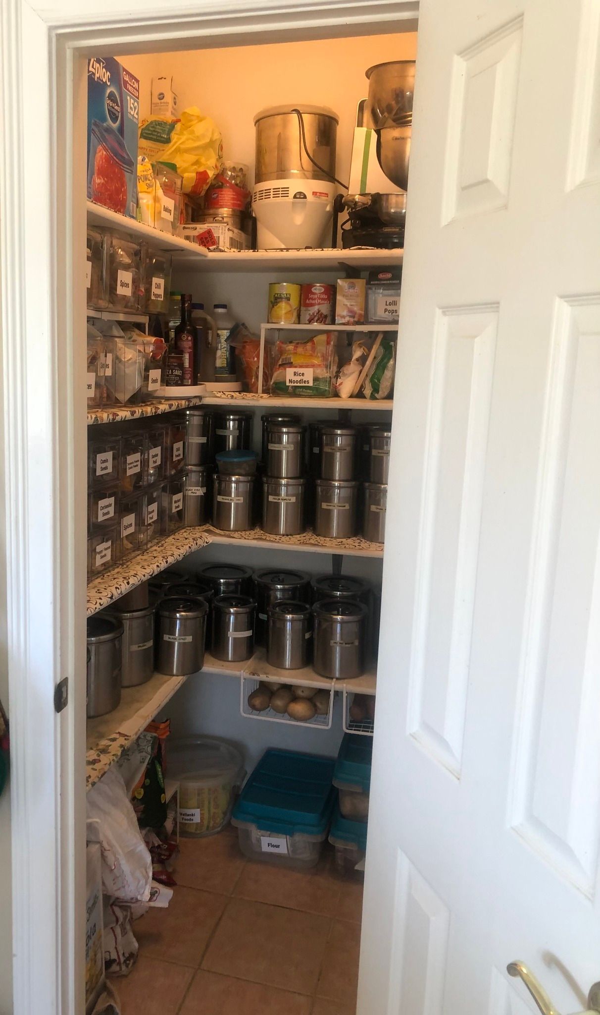Pantry with shelves full of food items and spices in silver containers, viewed through an open white door.