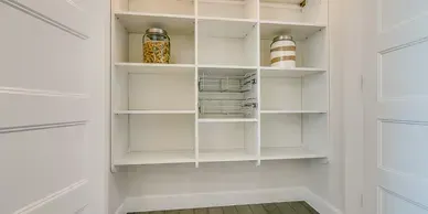 White built-in pantry with multiple shelves. Two decorative jars sit on top shelf. Wooden floor and white door frames.