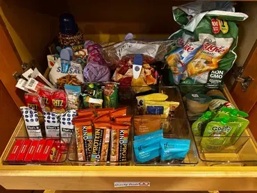 Organized pantry drawer with snacks: chips, crackers, protein bars, and pouches in clear containers.