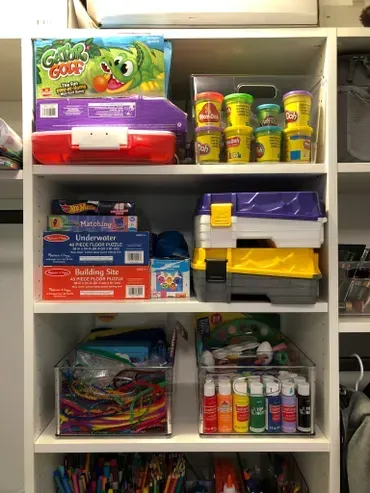 White closet shelves with toys and craft supplies stored in bins and boxes.