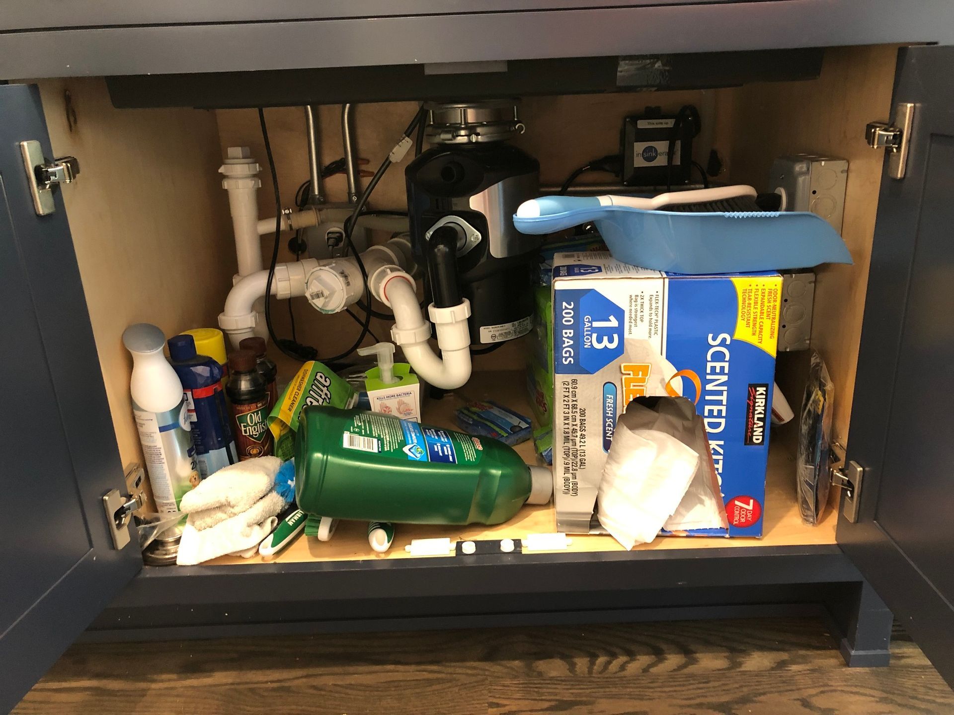 Under-sink cabinet with plumbing, garbage disposal, cleaning supplies, and trash bags.