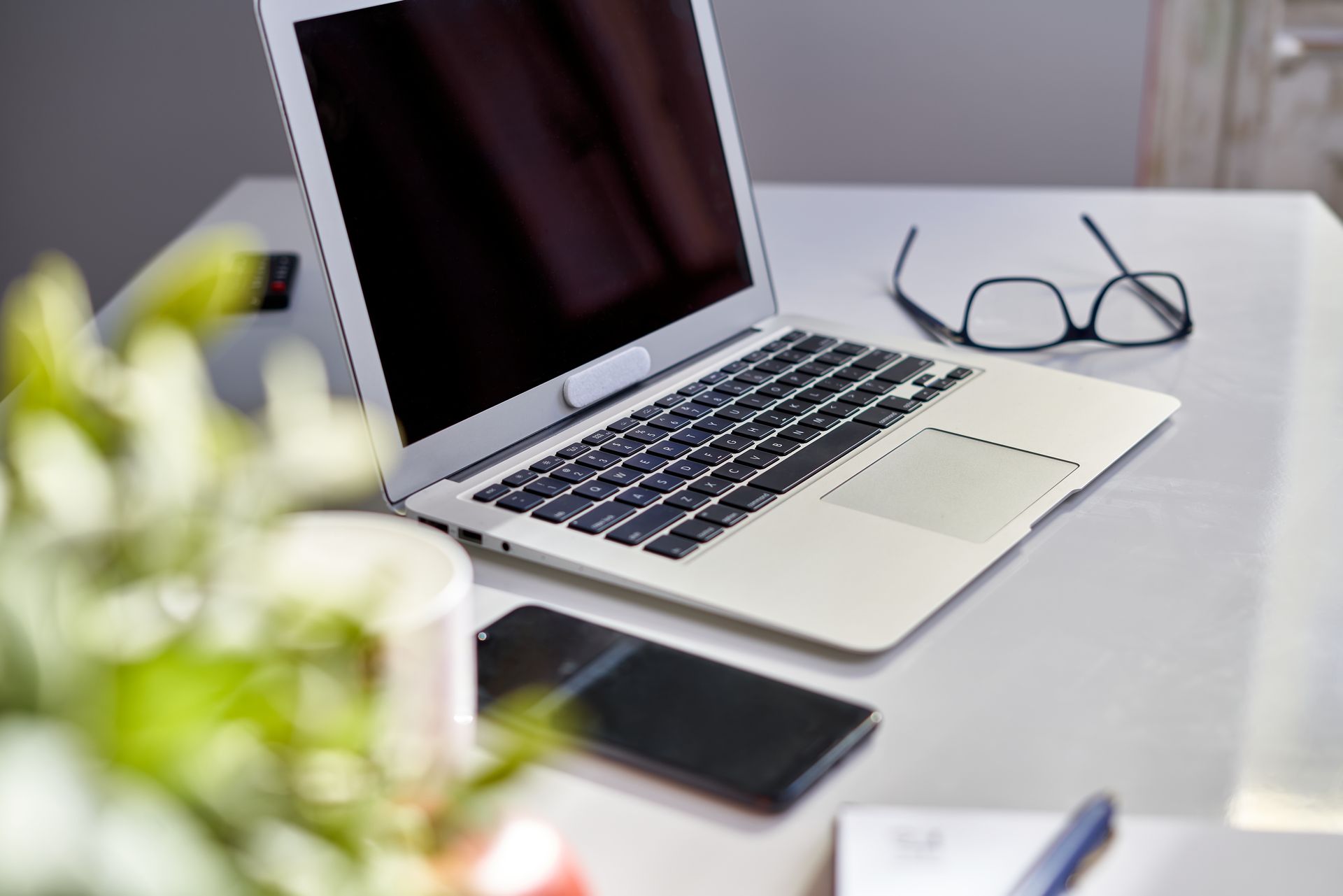 A laptop, smartphone, and glasses sit on a desk next to a blurred plant.
