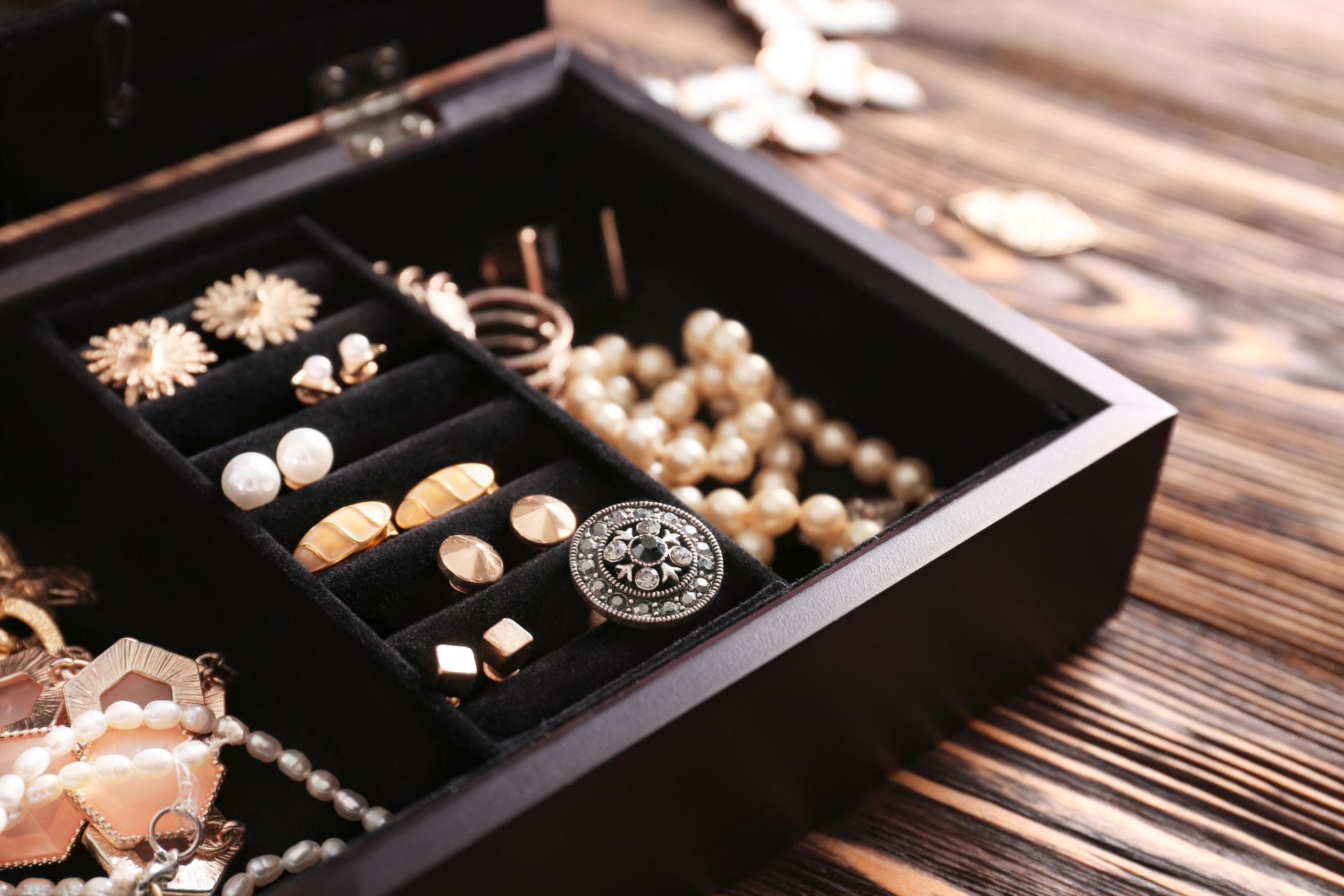 An open, black wooden jewelry box sits on a dark, rustic wooden table, holding pearls, rings, and various earrings.
