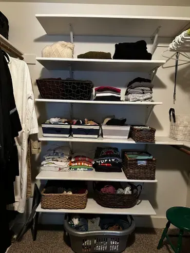 Closet with white shelves holding folded clothes, baskets, and a laundry hamper on the floor.