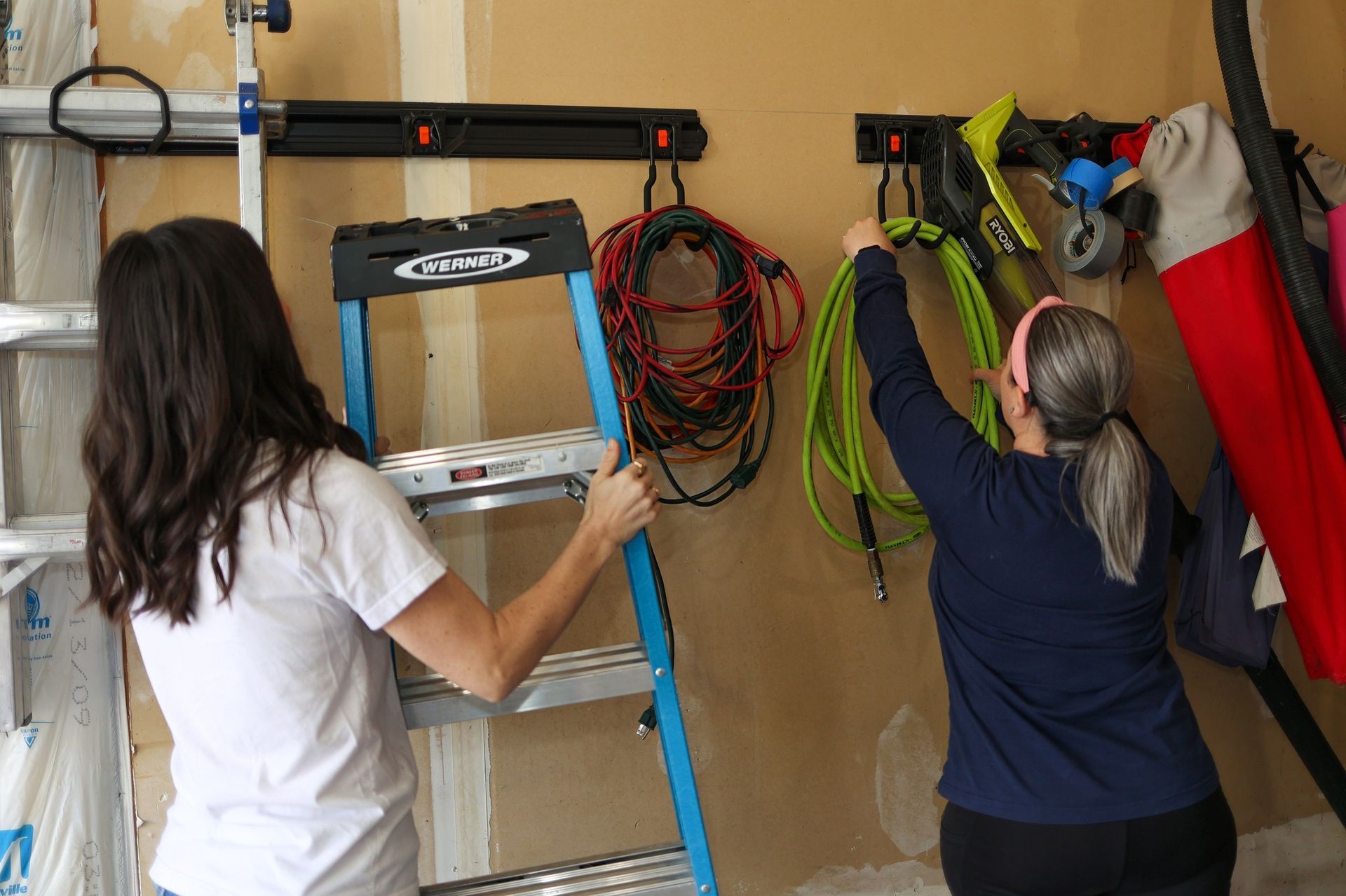 Two people organizing items on hooks in a garage. One holds a ladder, the other hangs a hose.