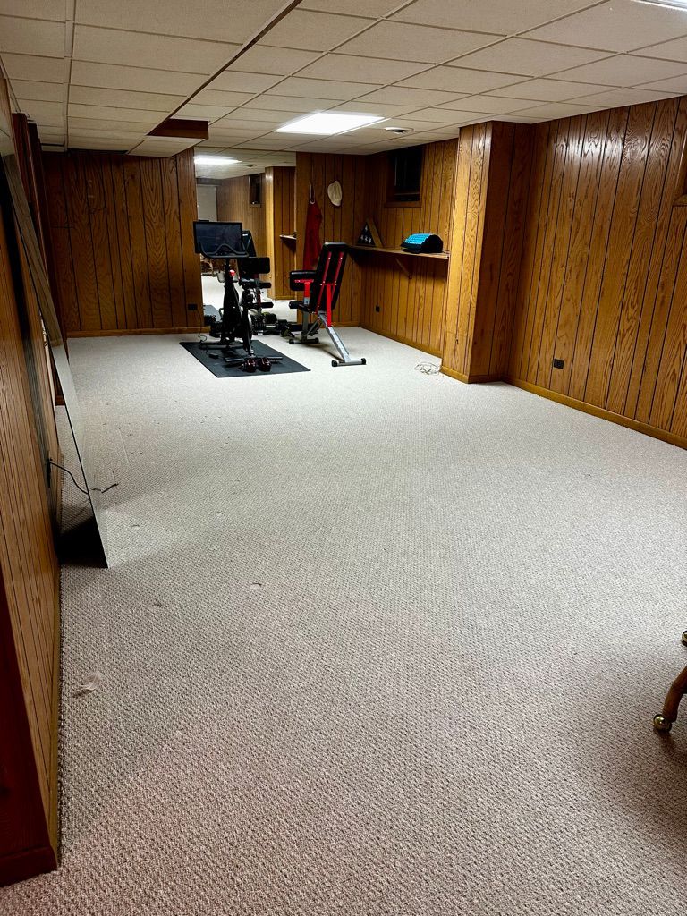 A carpeted basement room with exercise equipment. Wooden paneling lines the walls and ceiling.