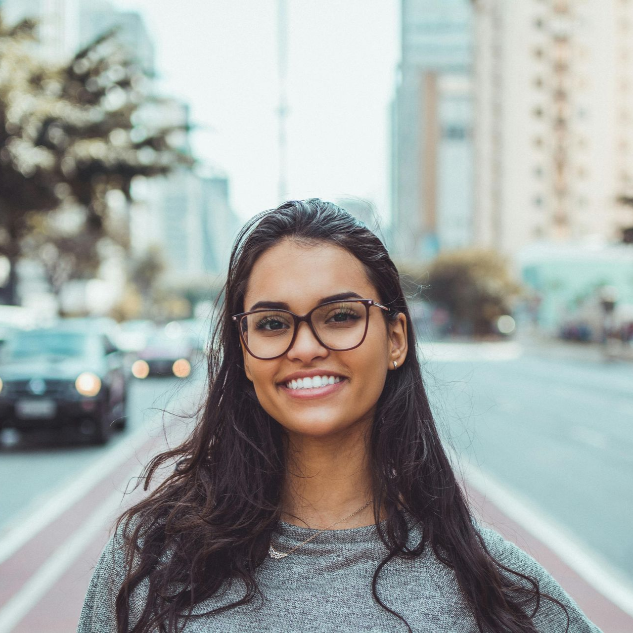 Woman with glasses smiling in a city street, cars blurred in the background.