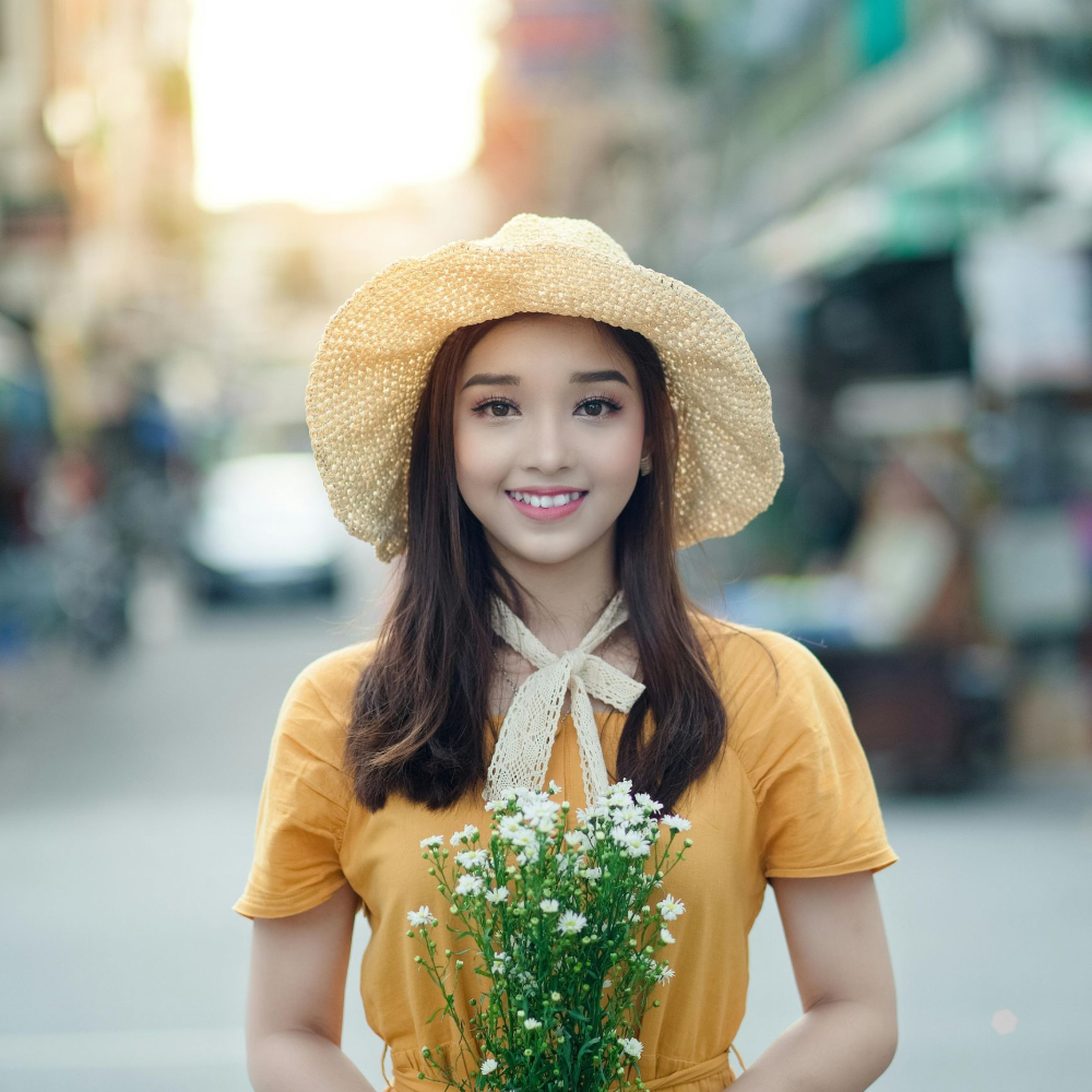 Woman wearing a yellow dress and straw hat, holding flowers, smiling, with a street background.