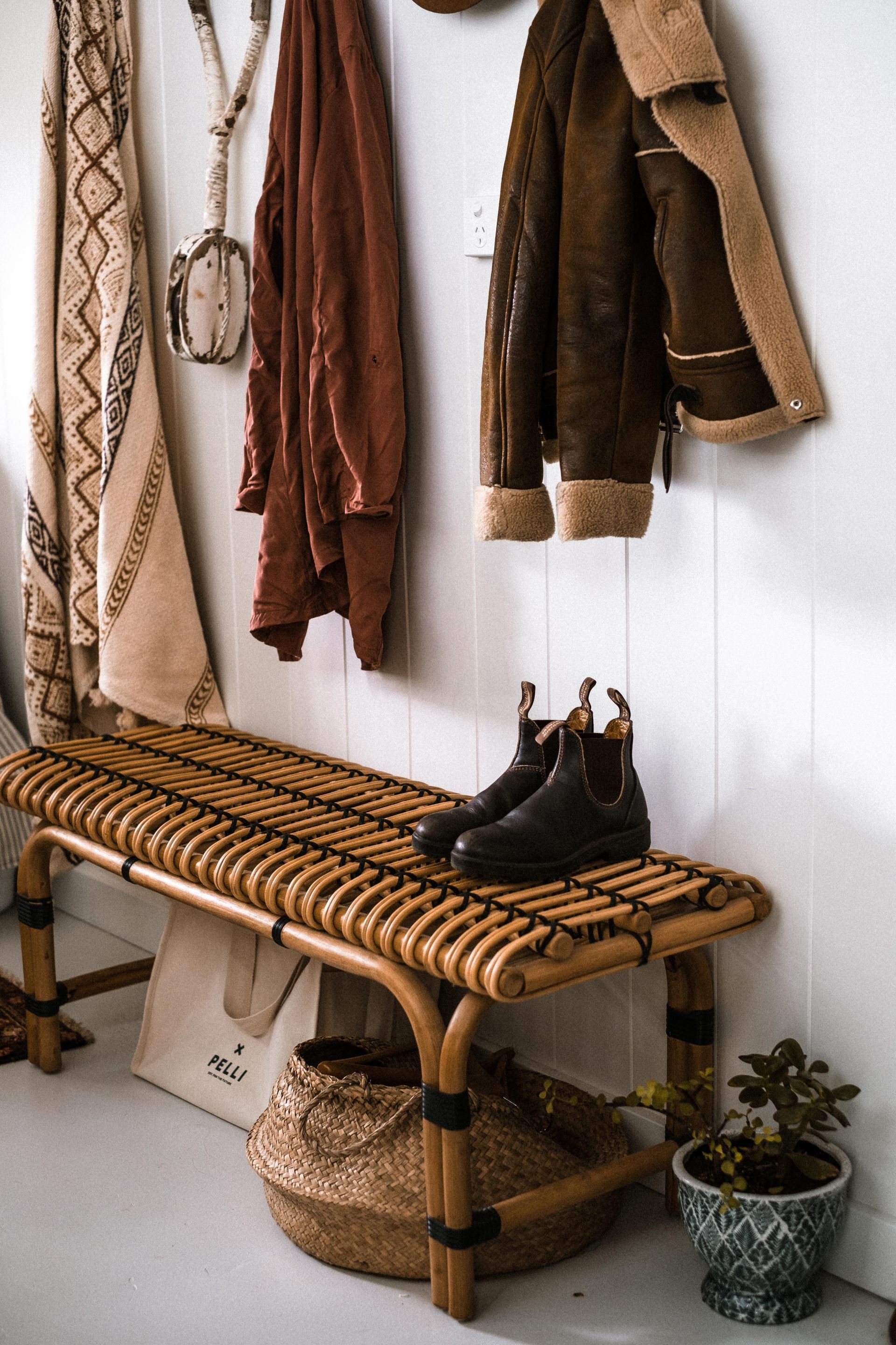 Cozy entryway bench with hanging coats, boots, basket, and small potted plant against a white wall.