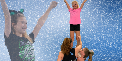 Cheerleaders in action: two are lifting a girl in a pink shirt, another smiles with arms raised, glittery blue background.