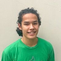 Young man with curly brown hair in a green shirt smiles. Gray background.