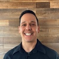 Man smiling, wearing a dark shirt, in front of a wooden wall.