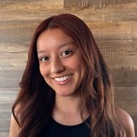 Woman with auburn hair smiles in front of a wooden wall, wearing a black top.