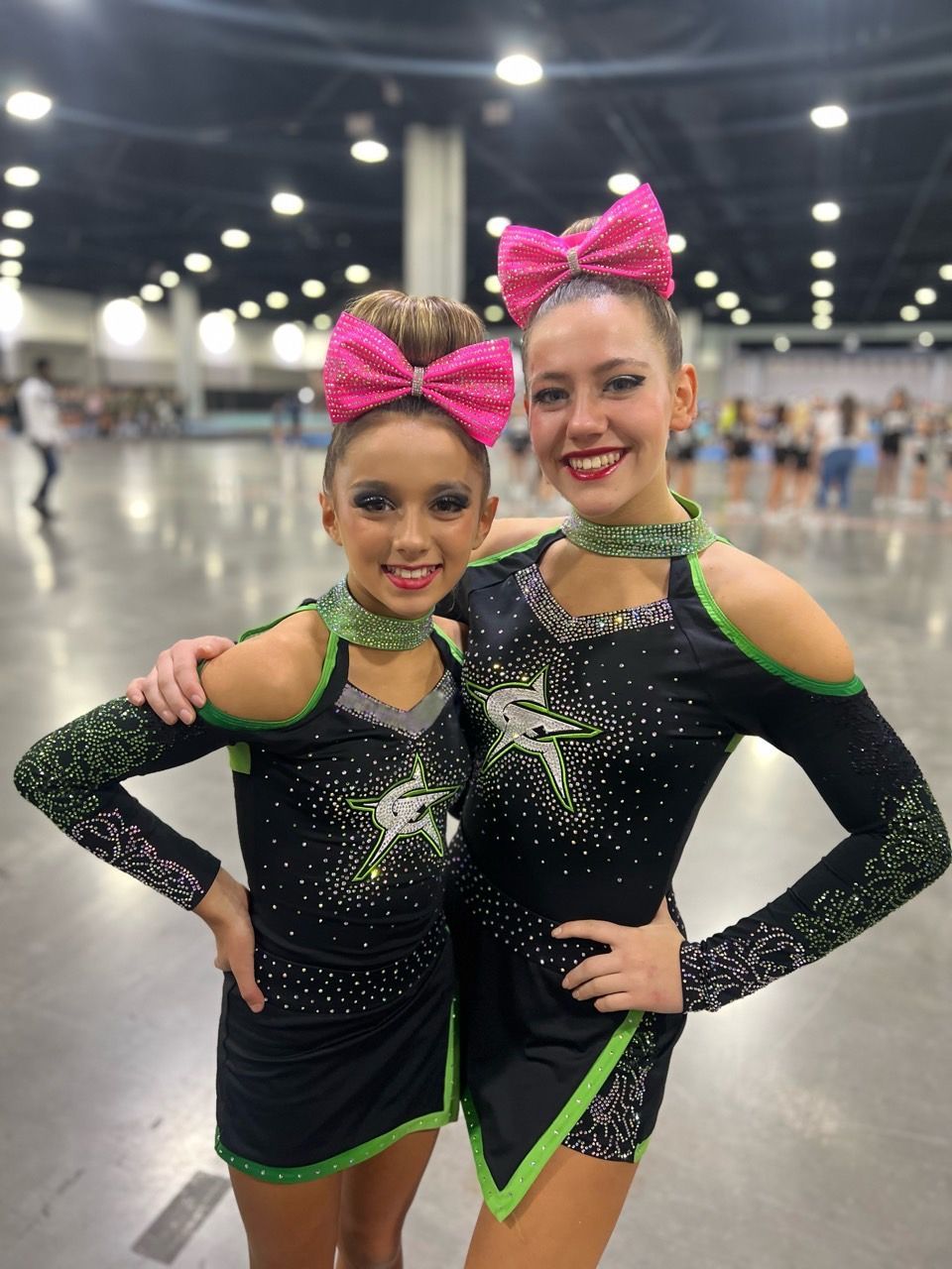 Two cheerleaders in black and green uniforms pose together at a competition, smiling, with pink bows.
