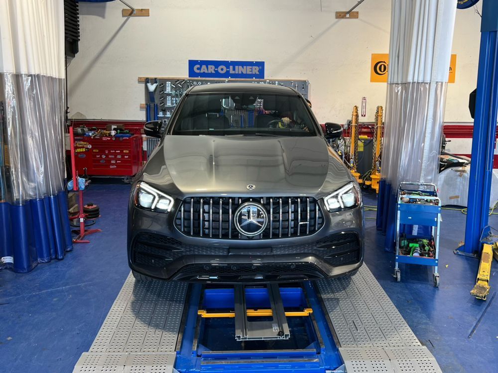 Dark gray Mercedes SUV in a repair shop, positioned on a lift. Shop interior has tools, equipment, and a Car-O-Liner sign. | Ma's Auto Body