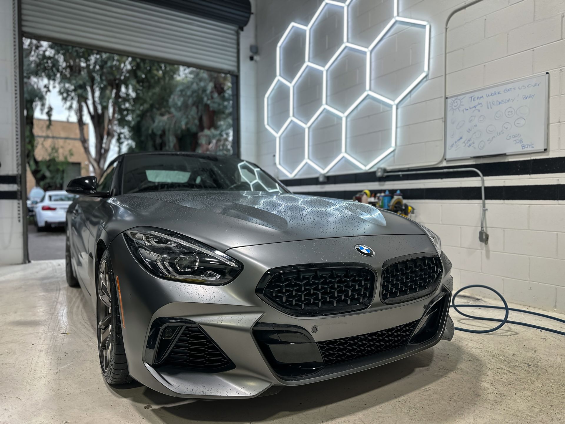 Gray BMW Z4 roadster in a car detailing shop, parked beneath a honeycomb-shaped neon light fixture. | Ma's Auto Body