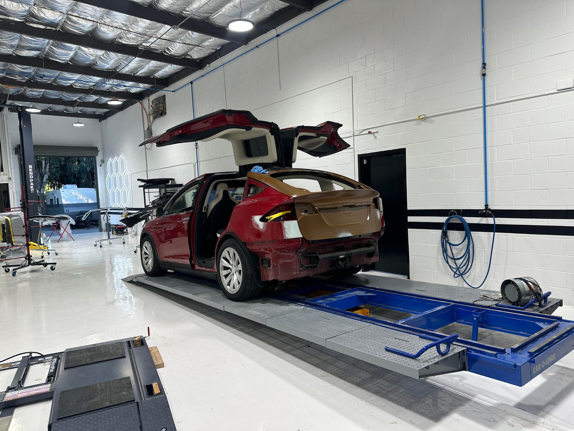 Red Tesla SUV in auto shop, doors open. Rear and side panels removed, raised on platform.| Ma's Auto Body