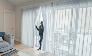 A woman is opening a sliding glass door with white curtains.